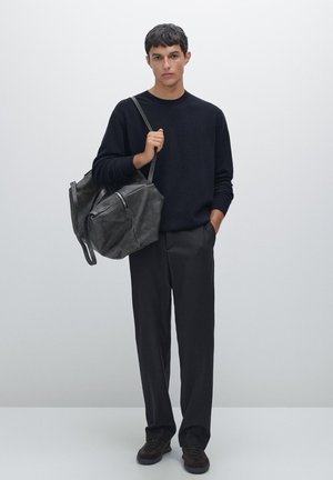 Zwarte gebreide trui, donkergrijze broek, houdt een grote zwarte leren tas vast met een rits en banden. Eenvoudig, gestructureerd ontwerp, matte afwerking.