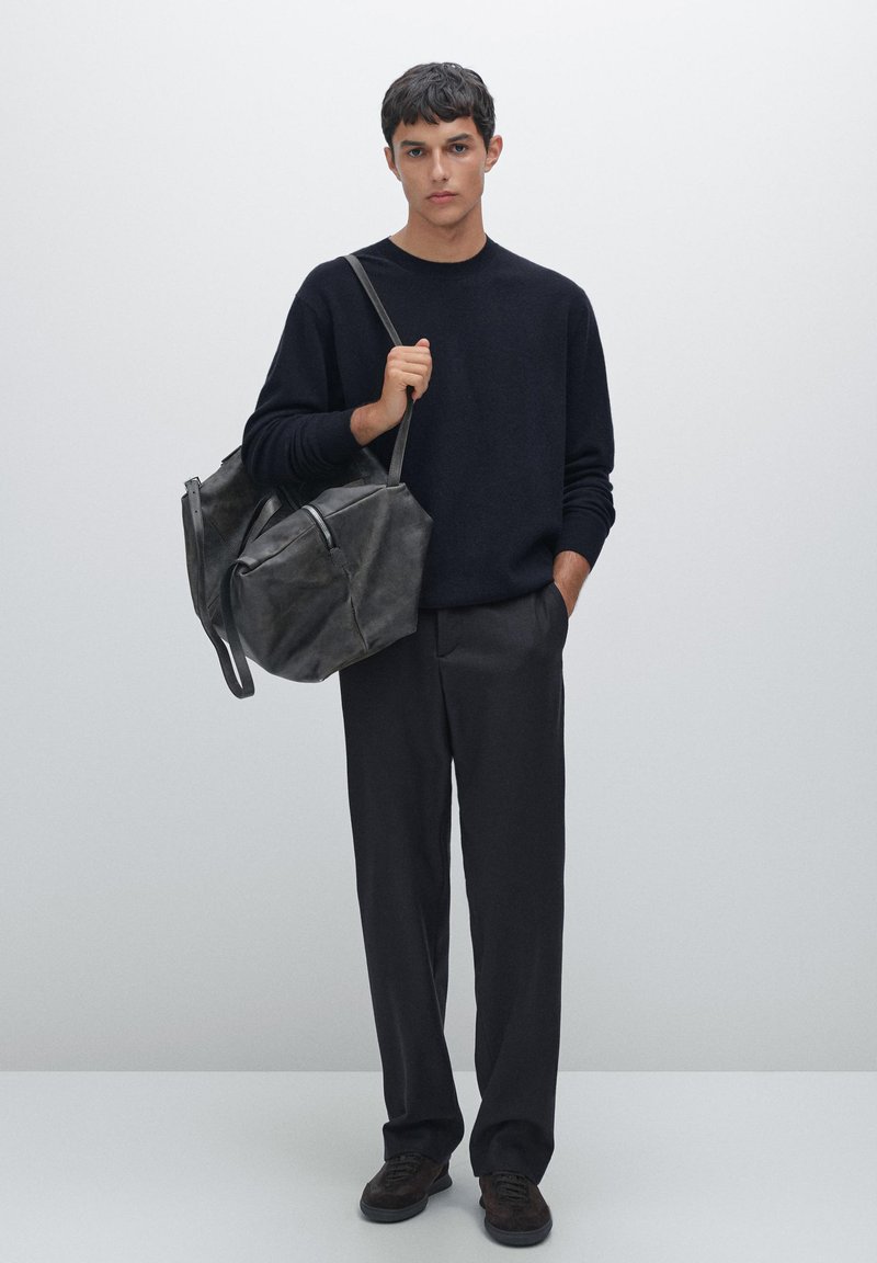 Schwarzer Strickpullover, dunkelgraue Hose, hält eine große schwarze Lederhandtasche mit Reißverschluss und Riemen. Einfaches, strukturiertes Design, matte Oberfläche.