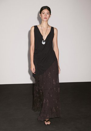 Schwarzes ärmelloses Kleid aus geschichtetem, strukturiertem dunklem Stoff. Besitzt einen tiefen V-Ausschnitt und ein drapiertes Design, ergänzt durch eine runde Anhänger-Halskette.