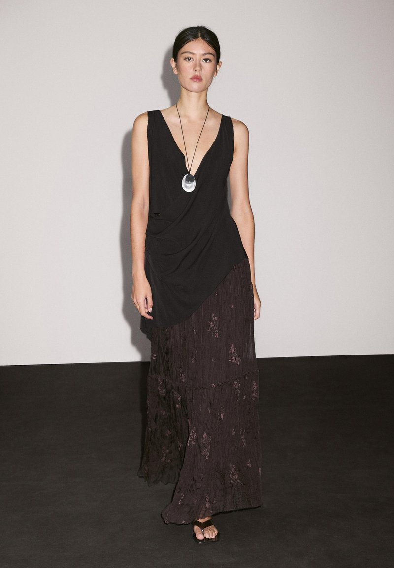 Black sleeveless dress with layered, textured dark fabric. Features a deep V-neck and draped design, accessorized with a round pendant necklace.