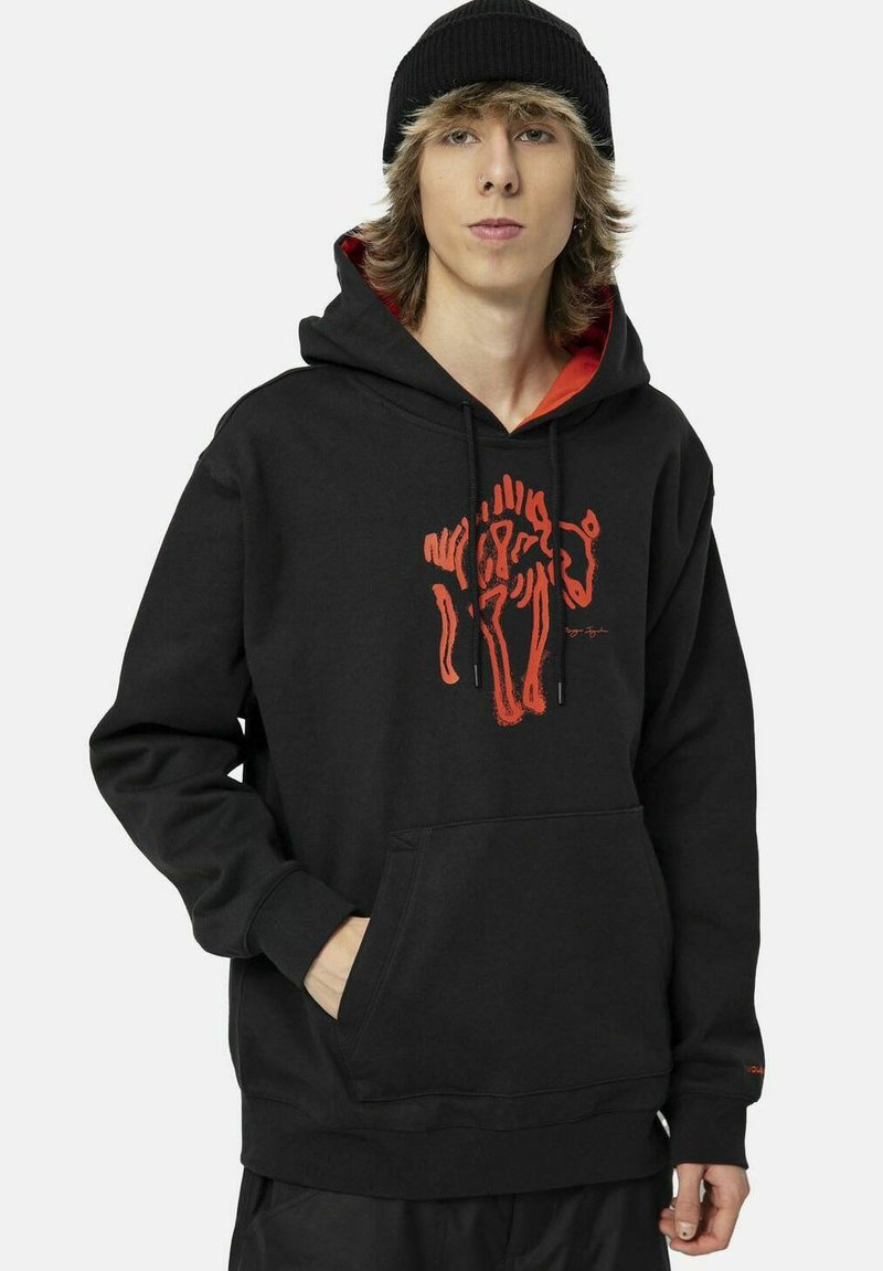 Jeune personne portant un bonnet noir et un sweat à capuche noir avec un graphique de lion orange et les mains dans la poche avant, debout devant un fond uni.
