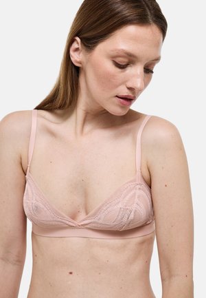 Femme portant un bralette en dentelle rose clair avec des bretelles fines et réglables, regardant vers le bas et légèrement à sa gauche sur un fond uni.