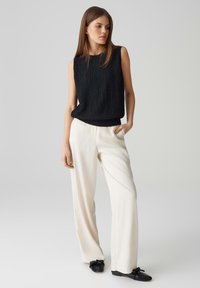 Top senza maniche di colore nero con texture, abbinato a pantaloni larghi di colore crema e scarpe slip-on nere. Design semplice con un forte contrasto di colori.