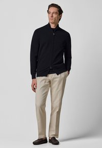 Czarny sweter na zamek z wysokim kołnierzem, zestawiony z beżowymi spodniami i ciemnobrązowymi butami. Gładka tekstura, dopasowany krój i minimalistyczny wygląd.