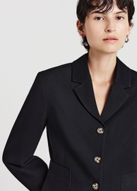 Blazer nero con rever a incavo, chiusura con quattro bottoni a contrasto e due tasche frontali. Tessuto liscio e vestibilità sartoriale.