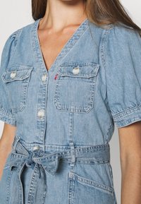 Vaaleansininen farkkumekko, jossa on V-pääntie, lyhyet puffihihat, kaksi rintataskua, helmiötäiset napit ja vyötärössä vyötyyli.