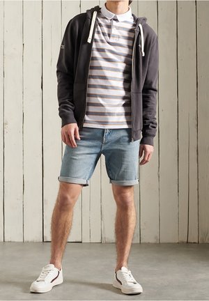 Mand iført stribet polo, mørk hættetrøje med lynlås, opkørte denimshorts og hvide sneakers, stående op ad en væg af træpaneler.
