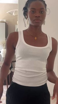 Top aderente bianco a costine, abbinato a pantaloni neri a vita alta. Collana dorata con pendente, capelli texturizzati, sfondo neutro.