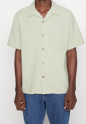 Mężczyzna w jasnozielonej koszuli na krótki rękaw z kieszenią na piersi i niebieskich dżinsach, stojący na białym tle.