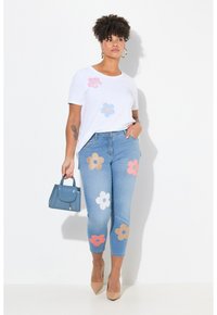 Camiseta blanca con apliques de flores coloridas, jeans de mezclilla azul claro con parches florales, sosteniendo un pequeño bolso azul, zapatos de tacón marrones.