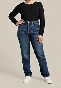 Person iført mørkeblå højtaljede jeans, sort langærmet top og hvide sneakers, stående med den ene hånd på hoften mod en ensfarvet baggrund.