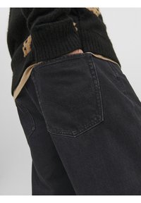 Pantalones de denim negro con un acabado texturizado, que cuentan con bolsillos traseros cosidos y un diseño ligeramente ajustado. Llevados con una camisa de color marrón grisáceo por debajo.