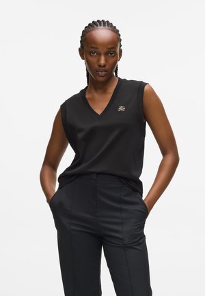 Haut noir sans manches avec un décolleté en V et un logo doré discret. Associé à un pantalon noir taille haute bien ajusté avec poches.