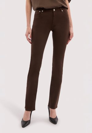 Mujer con pantalones ajustados de color marrón oscuro y tacones negros de punta, de pie contra un fondo claro y uniforme.