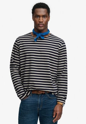 Hombre con camisa de manga larga a rayas blancas y negras, vaqueros azules, cinturón trenzado marrón, pañuelo azul al cuello y reloj de pulsera, de pie con una mano en el bolsillo.