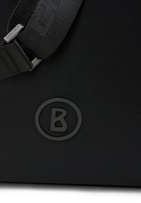 Svart tygväska med en strukturerad yta, en rund präglad logotyp med ett 'B', samt ett robust justerbart band med metallbeslag.