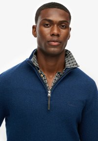 Suéter navy con cremallera y tejido texturizado, sobre una camisa a cuadros con cuello. El suéter tiene un logo sutil en el pecho.