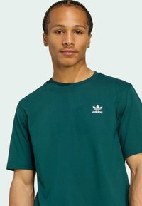 Zelené bavlnené tričko s okrúhlym výstrihom, krátkymi rukávmi a bielym logom Adidas na ľavej hrudi. Rovnomerná textúra a klasický strih.