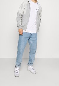 Sudadera gris con cremallera y mangas gris claro, camiseta blanca debajo, jeans azules y zapatillas blancas. Cuenta con un bolsillo frontal y capucha ajustable.