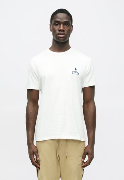 Νεαρός άνδρας φορώντας λευκό μπλουζάκι Polo Ralph Lauren και μπεζ cargo παντελόνι, στέκεται μπροστά σε απλό ανοιχτό φόντο, κοιτώντας μπροστά.