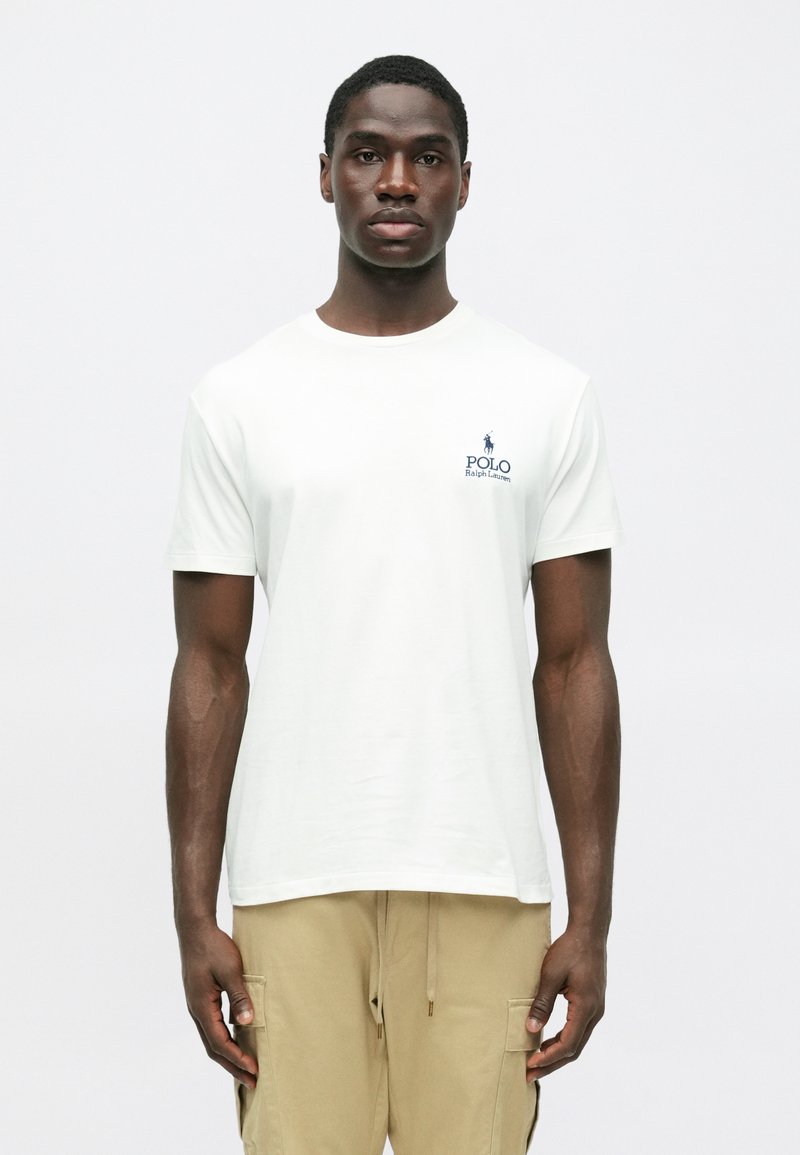 Νεαρός άνδρας φορώντας λευκό μπλουζάκι Polo Ralph Lauren και μπεζ cargo παντελόνι, στέκεται μπροστά σε απλό ανοιχτό φόντο, κοιτώντας μπροστά.