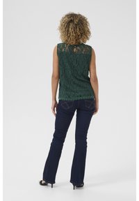 Top sans manches en dentelle vert foncé avec un ourlet festonné et un dos à ouverture en clé, associé à un jean évasé bleu foncé et des talons noirs.