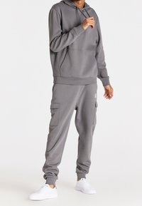 Šedá mikina s kapucí a kalhoty typu cargo vyrobené z měkké tkaniny, s přední kapsou, zúženými nohavicemi a elastickými manžetami. Doplněno bílými teniskami.