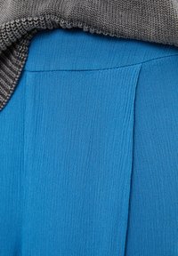 Sininen teksturoitu kangas laskeutuu harmaan neulotun reunan vierelle, jossa on sileä sauma, joka yhdistää kaksi materiaalia.