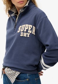 Granatowy polarowy sweter z białym haftowanym napisem "SUPER DRY", zamek w kołnierzu i białe paski na rękawach. Założony na pasiasty T-shirt.
