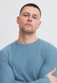 Pull bleu clair en tissu doux, avec un col rond et des manches longues ajustées, porté par une personne les bras croisés.