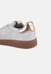 Zapatilla de cuero perforada blanca con un tacón metálico dorado y suela de goma marrón. Presenta un logotipo en el tacón y acentos texturizados.