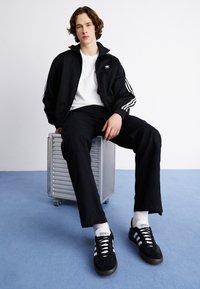 Young man with curly hair wearing black Adidas jacket, white t-shirt, black pants, and sneakers sitting on a metal box on blue carpet.
