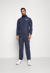 Navy tracksuit with a zip-up jacket and elastic waistband pants. Features a logo on the chest, sporty design, and white sneakers.