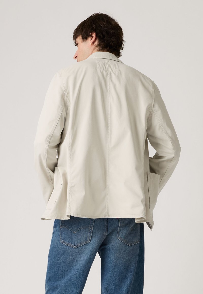 Homme portant une veste beige clair et un jean bleu, vu de dos sur un fond clair uni.