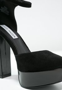 Chaussure à plateforme noire à talons hauts en daim avec une finition en cuir verni brillant. Elle présente un talon épais et une large bride à la cheville.