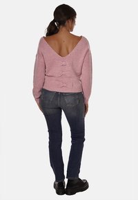 Maglione rosa a maglia con scollatura a V profonda, decorato con tre fiocchi lungo la spina dorsale. Abbinato a jeans blu scuro e stivaletti neri.
