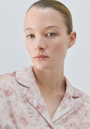Jeune femme aux cheveux tirés en arrière, portant un haut de pyjama floral rose clair, expression neutre, sur un fond clair uni.