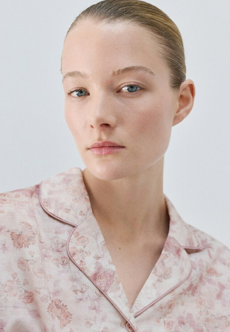 Mladá žena s učesanými vlasmi dozadu, oblečená v svetloružovej pyžamovej blúzke s kvetinovým vzorom, s neutrálnym výrazom, na jednoduchom svetlom pozadí.