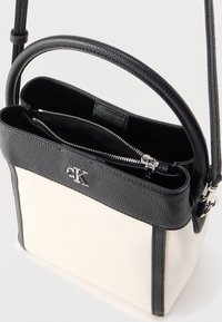 Μικρή ασπρόμαυρη τσάντα χιαστί με λεπτομέρειες από δέρμα με υφή, ασημένιο φερμουάρ και λογότυπο "cK" στην πάνω μπροστινή άκρη.