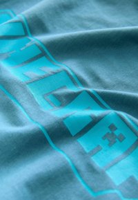 Teal bomulds T-shirt med en glat tekstur, prydet med markante turkise grafikker og et pixeliseret design, der viser folder og krøller.