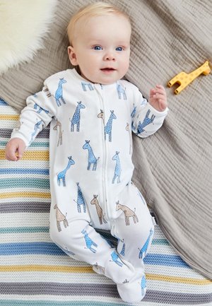 Weiße Reißverschluss-Jumpsuit mit blau-braunen Giraffenmustern, aus weichem Stoff, begleitet von einem gelben Giraffen-Spielzeug auf einer gestreiften Decke.