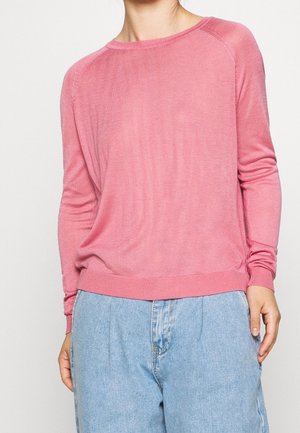 Kvinde iført en blød lyserød langærmet sweater kombineret med løse lyseblå denimjeans, mod en ensfarvet lys baggrund.