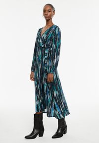 Vestido largo y fluido en negro con patrones abstractos en teal y blanco. Presenta un escote en V, mangas largas y una cintura atada. Combinado con botas negras hasta el tobillo.