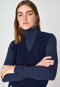 Marineblaues Rollkragenoberteil unter einem ärmellosen V-Ausschnitt-Pullover mit einem gesprenkelten blau-roten Muster, geripptem Textil und verschränkten Armen.