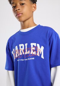 Blue cotton T-shirt featuring white "HARLEM" text with red accents, short sleeves, and a crew neckline, layered over a white long-sleeve shirt.
