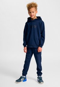 Granatowy komplet z bluzy z kapturem i spodni dresowych wykonany z miękkiego materiału, z logo na środku i prążkowanymi ściągaczami. Buty sportowe z niebieskimi akcentami.