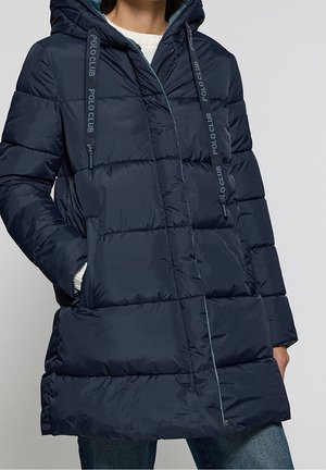 Femme portant une doudoune matelassée à capuche bleu marine avec des cordons imprimés "Polo Club" et les mains dans les poches latérales.