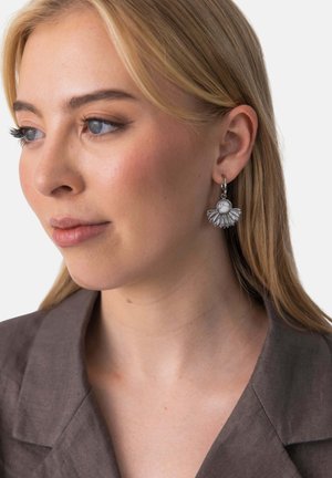 Blonde Frau in braunem Blazer trägt silbernen fächerförmigen Ohrring mit weißem Stein und blickt leicht nach links vor schlichtem Hintergrund.