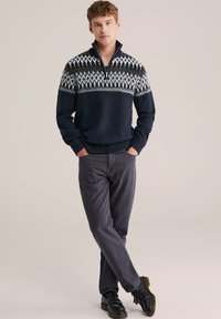 Pulover tricotat bleumarin cu guler cu fermoar și model geometric alb. Asortat cu pantaloni gri și pantofi negri. Textură netedă, design casual.