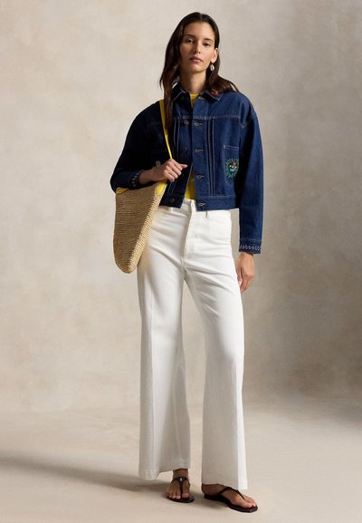 Femeie purtând o geacă din denim închis la culoare, pantaloni albi largi, sandale negre și având o geantă mare țesută pe umăr, pe un fundal simplu.